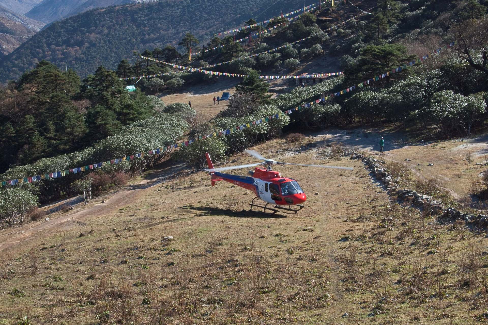 A helicopter on the Tengboche landing ground, rotor turning, prayer flags across the slope.