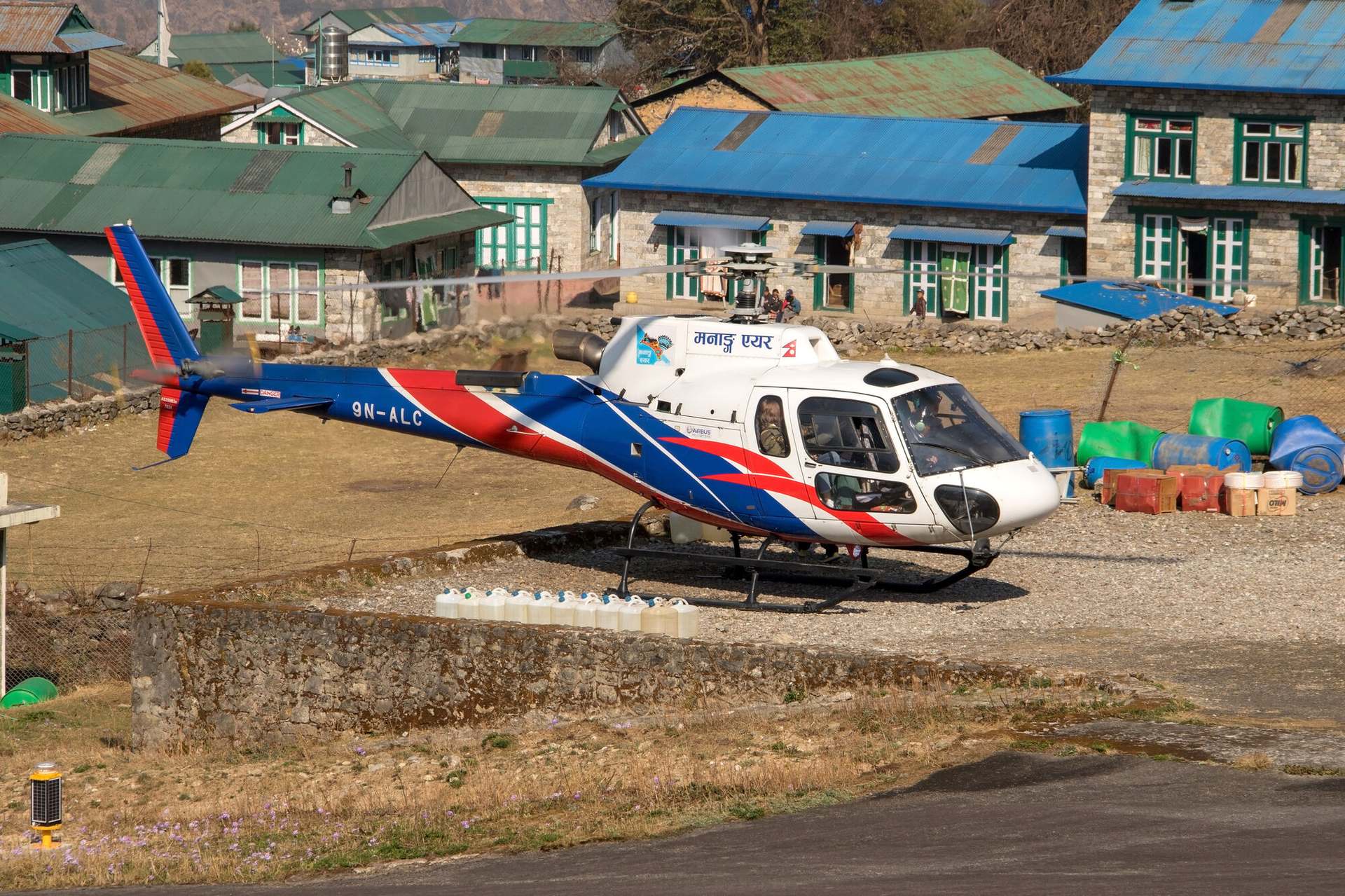 A single-engine charter helicopter on the pad at Lukla, the village and its terraces rising behind.