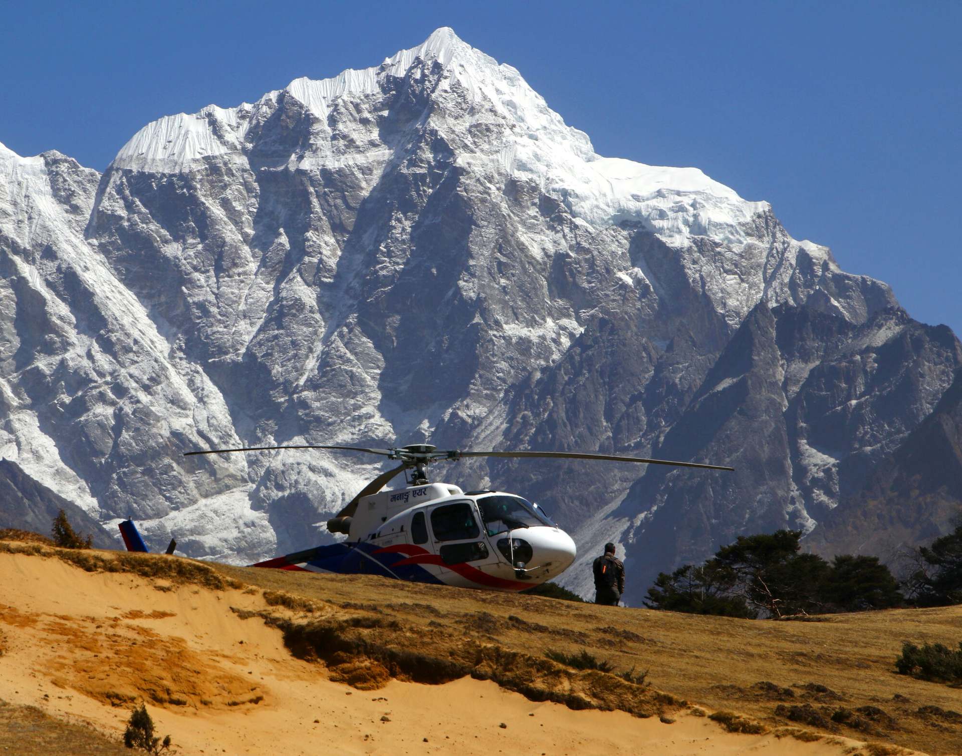 A helicopter on a hillside landing ground above Namche, the face of Taboche filling the sky behind it.