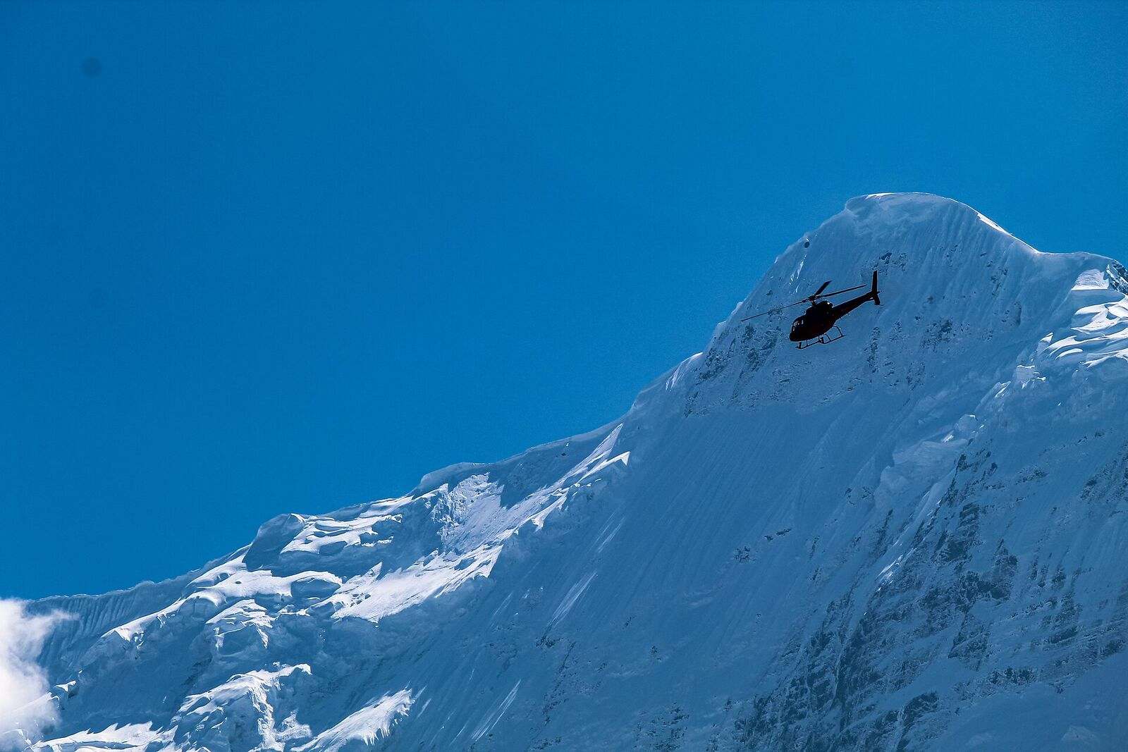 A helicopter working a high ridge line, small against the snow face behind it.