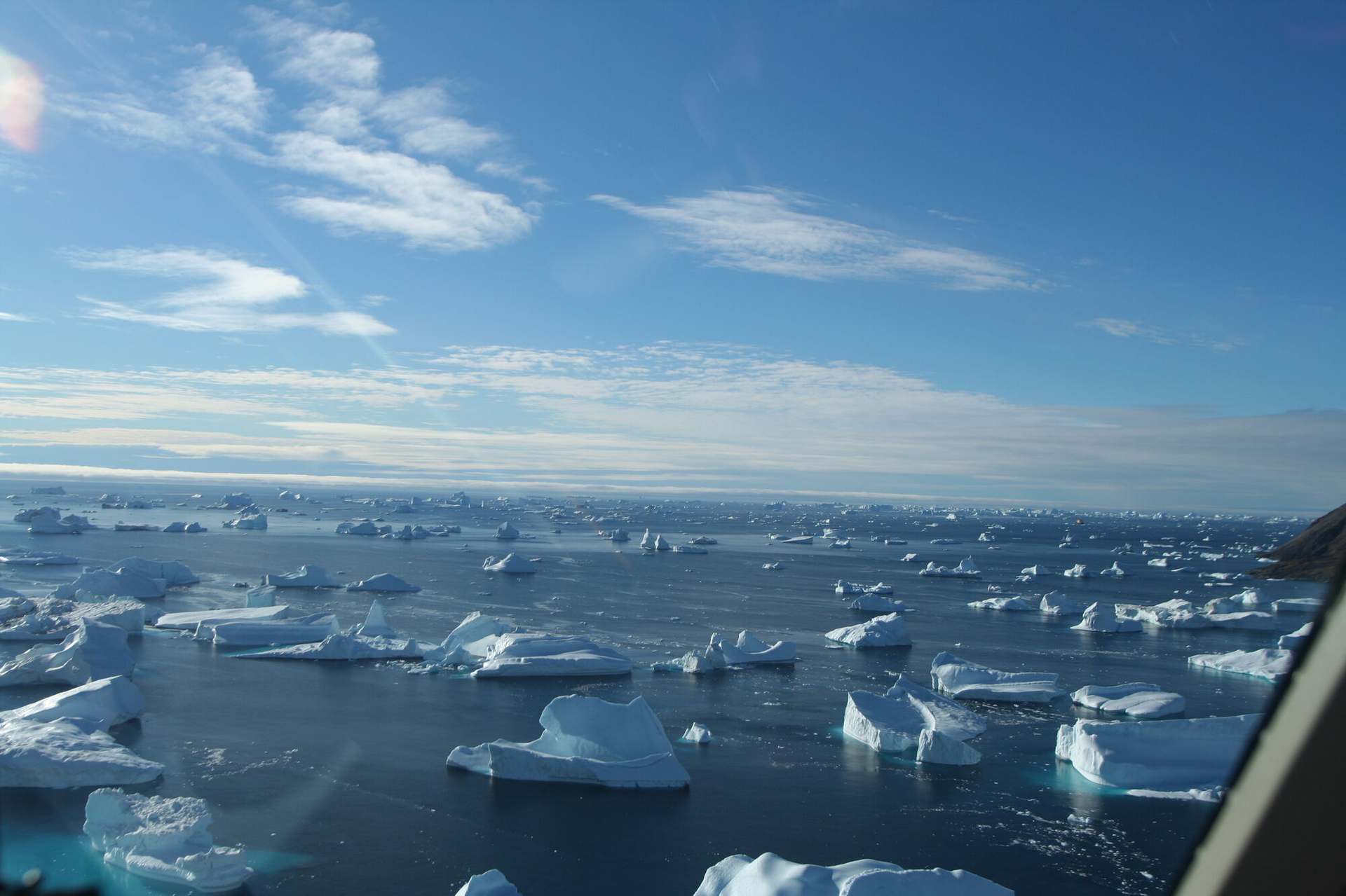 Ice and open water passing below, seen forward through the windscreen of a helicopter from the left-hand seat.