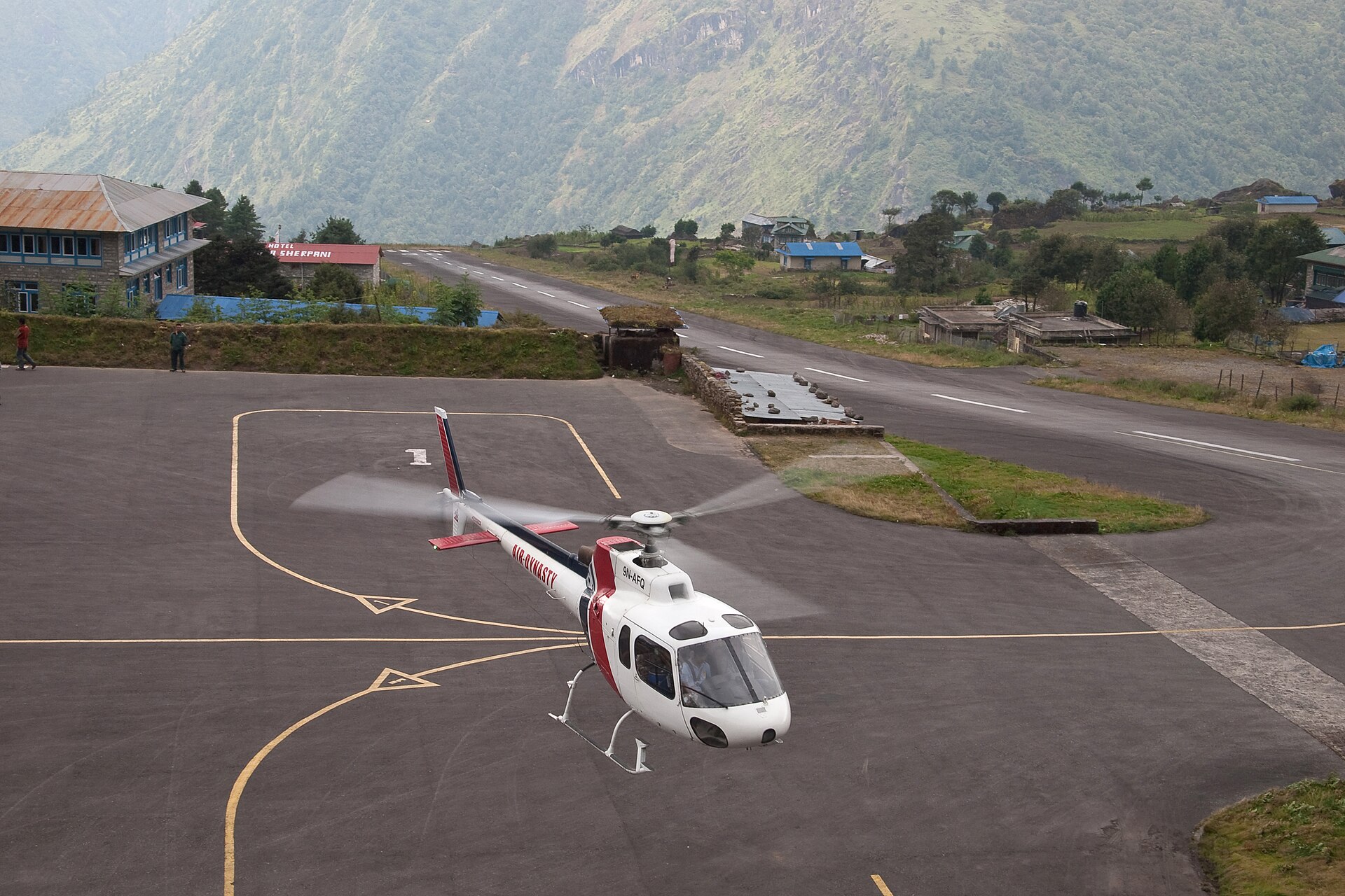 Lukla's sloping runway seen from above the apron, with a helicopter lifting off the marked pad in front of it.